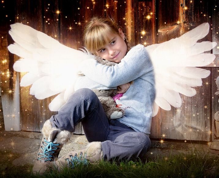 A young smiling girl with white angelic wings surrounded with sparkles of light sitting on the ground hugging a stuffed toy