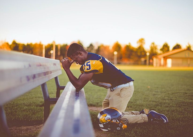 A black football player dressed in a blue and yellow outfit kneeling and praying with elbows on a bench