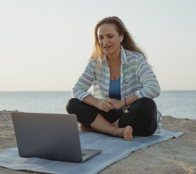 long-haired woman sitting cross-legged on a beach watching a movie on the laptop