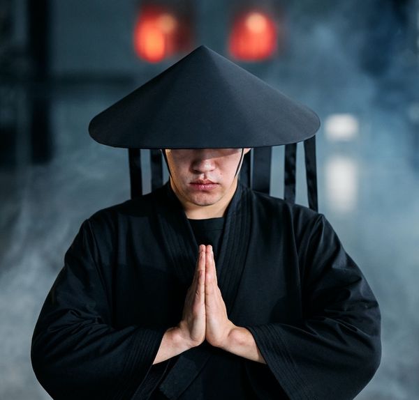Asiatic man dressed in black suit and a cone shaped hat joining hands in prayer