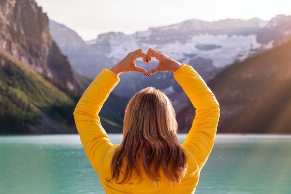 back view of-woman in yellow long sleeve shirt with heart hand gesture in front of a mountain lake