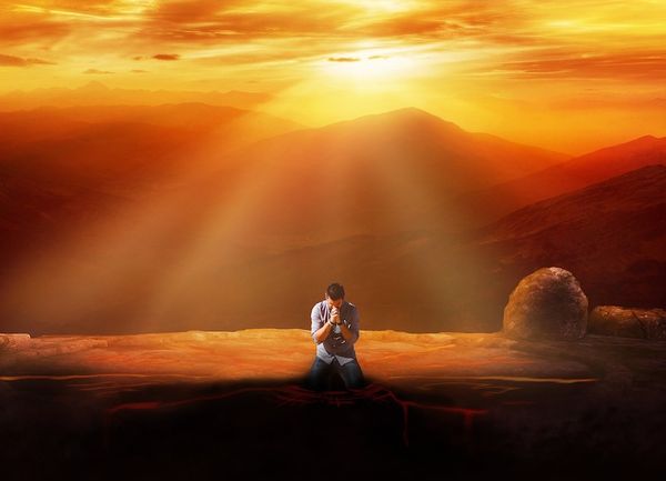 A man kneeling down in the act of praying illuminated by rays of the sun in a fantasy red landscape